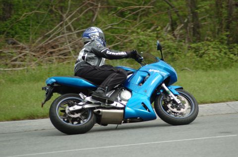 2007 Kawasaki Ninja R - A rider on a blue motorcycle cruises down a road, with trees and foliage in the background. The rider's jacket displays the Kawasaki logo.
