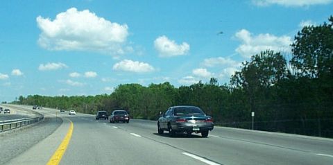 2001 Ford Crown Victoria - A dark-colored sedan travels down a multi-lane highway under a partly cloudy sky.