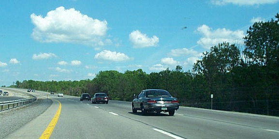 Dark sedan driving on a highway with trees and clouds in the background