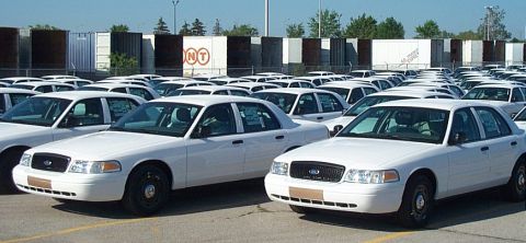 2001 Ford Crown Victoria - Large fleet lot packed with identical white Ford Crown Victoria Police Interceptor sedans on black steel wheels with protective tape strips…