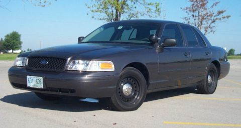 2001 Ford Crown Victoria - Grey Ford Crown Victoria Police Interceptor parked in an empty striped parking lot, shown from the front three-quarter angle with headlight…