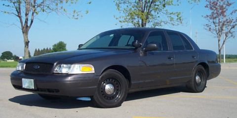 2001 Ford Crown Victoria - A dark gray Ford Crown Victoria Police Interceptor sedan photographed from the front three-quarter in an empty paved lot, wearing black ste…