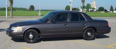 2001 Ford Crown Victoria - A gray/charcoal Ford Crown Victoria sedan shown in full side profile, parked in a paved lot with green fields, farm buildings and trees in…