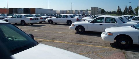 2001 Ford Crown Victoria - Rows of white Ford Crown Victoria Police Interceptor sedans with black steel wheels and window stickers sit in a paved staging lot, most st…