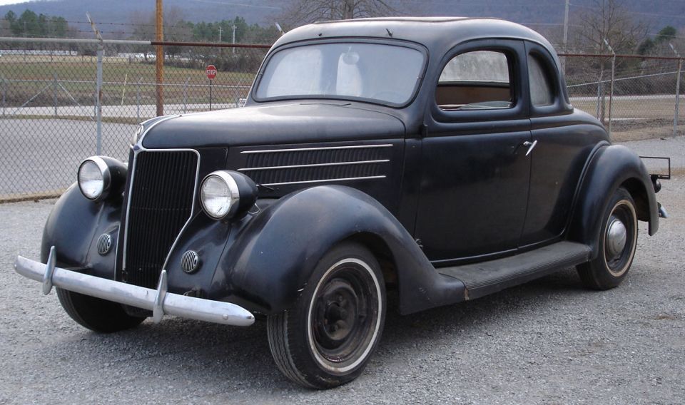 Weathered black 1936 Ford Coupe, front-left 3/4 view, on gravel by a chain-link fence.