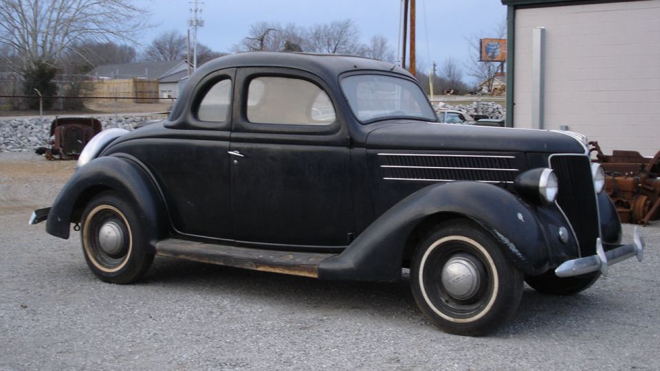 Black vintage 1936 Ford coupe, 3/4 front view on gravel with whitewall tires.