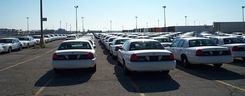 2001 Ford Crown Victoria - A large group of white 2001 Ford Crown Victoria police cars are parked in an outdoor lot under a clear sky.