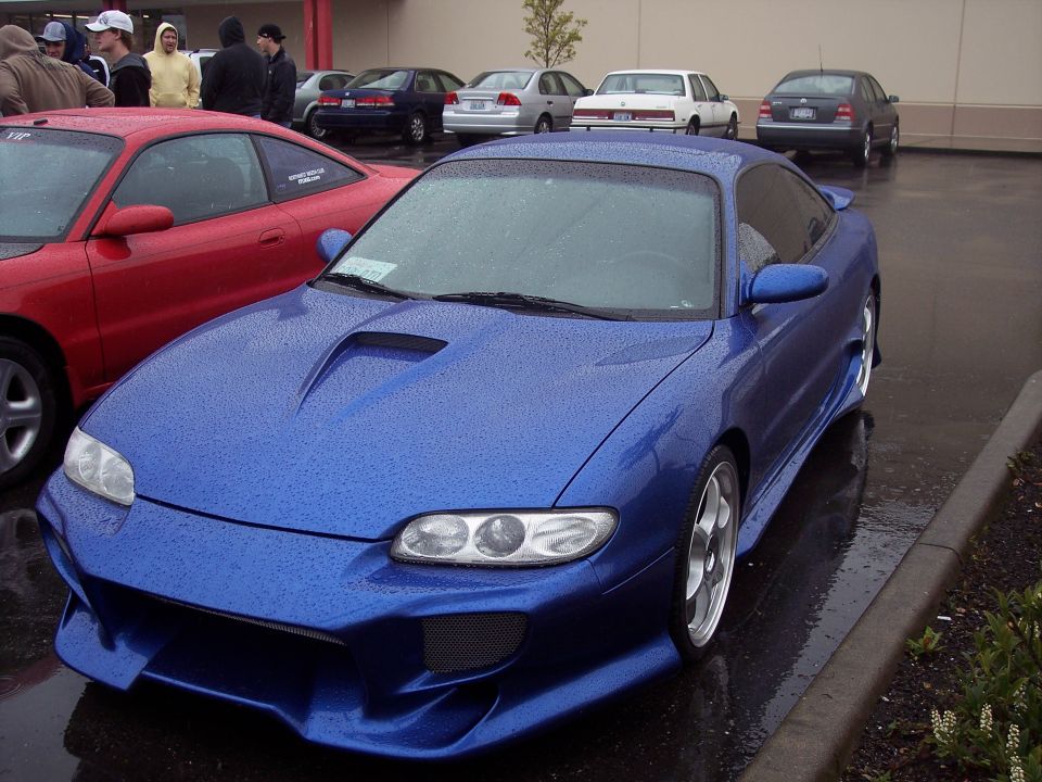 Blue modified coupe with body kit and hood scoop parked on a wet lot.