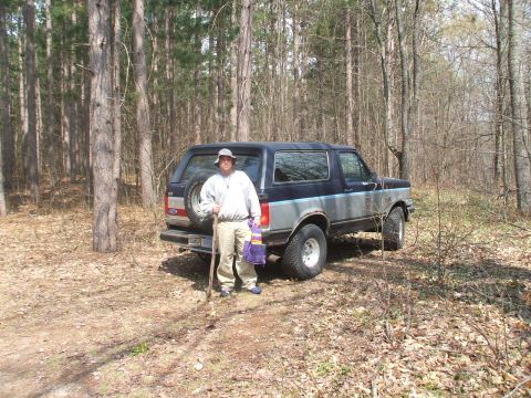 1990 Ford Bronco - A man stands next to a dark blue and gray Ford Bronco in a wooded area. He is holding a stick and a purple bag.