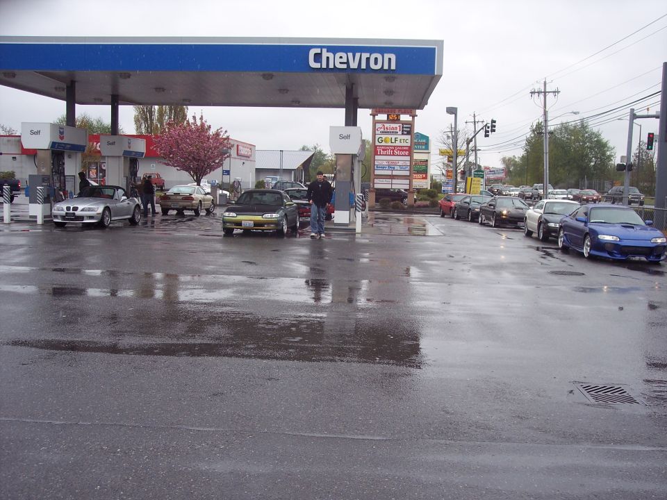 Blue coupe with racing stripes at a wet Chevron gas station.