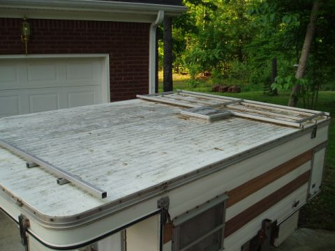 1980 Ford Bronco - The roof of a pop-up camper is shown, with a metal rack assembly on top. The camper is parked in front of a brick garage with a white door.