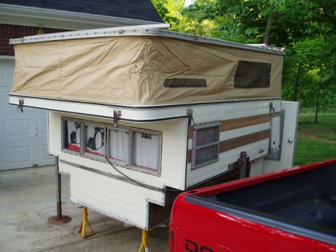 1980 Ford Bronco - A pop-up truck camper is shown set up on jack stands in front of a garage. The camper has a tan canvas pop-top and white siding with brown…