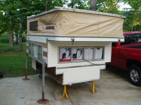 1980 Ford Bronco - A pop-up truck camper is shown supported by jack stands and stabilizers in a driveway. The camper has a tan canvas pop-top and a white body…
