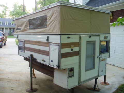 1980 Ford Bronco - A pop-up truck camper is shown supported by jacks in a driveway. The camper has a tan canvas pop-top section and white siding with brown wo…
