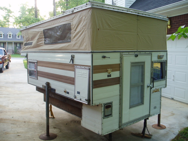 Pop-up truck camper with tan canvas top and white siding with brown trim, supported by jacks.