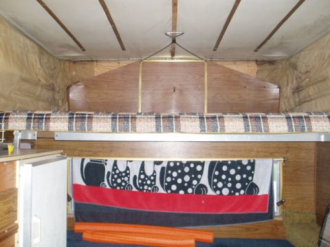 1980 Ford Bronco - Interior view of a pop-up camper bed with a plaid mattress and a decorative towel hanging below. The wooden interior features a refrigerato…