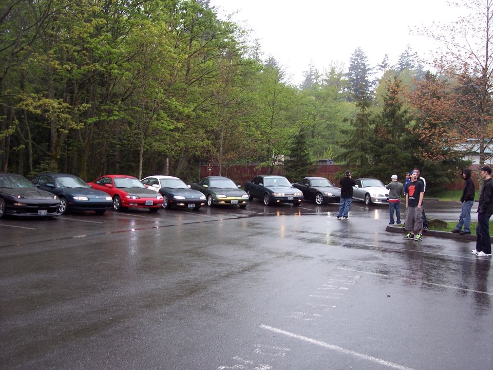 A line of sports cars parked in a wet lot with people standing around.