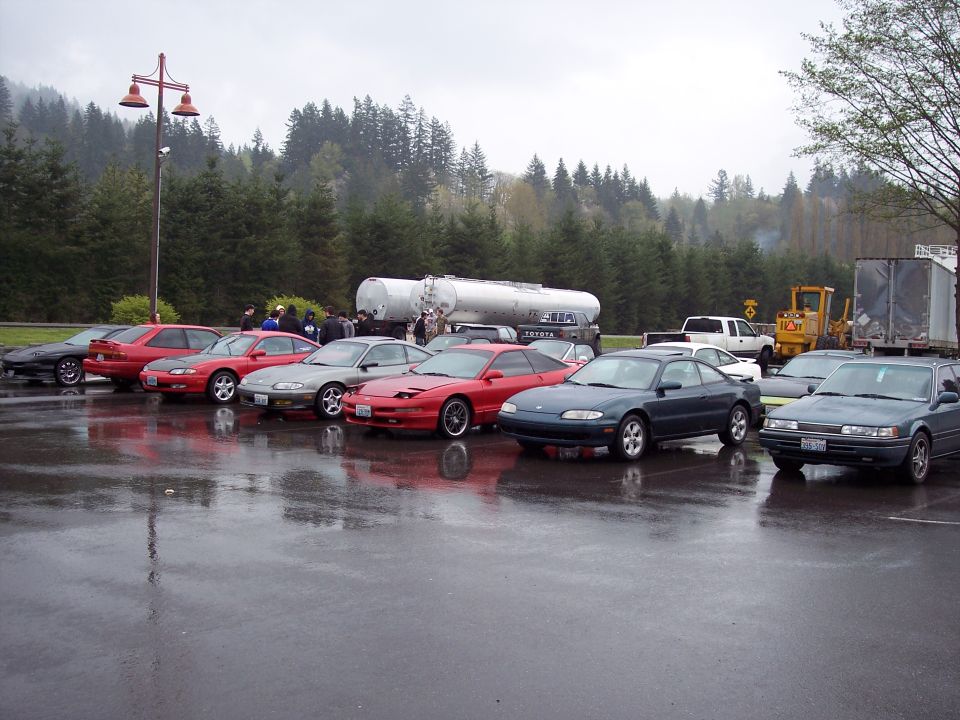 Several cars parked on a wet lot with trees and a tanker truck in the background.
