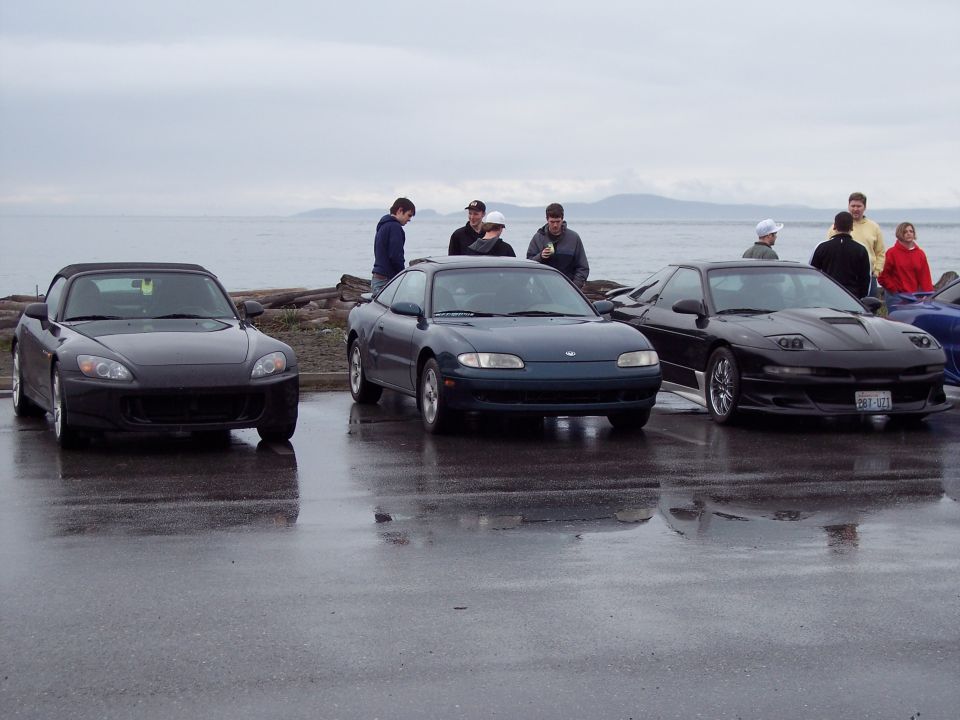 Black 1995 Ford Probe parked on wet asphalt with people and other cars nearby.
