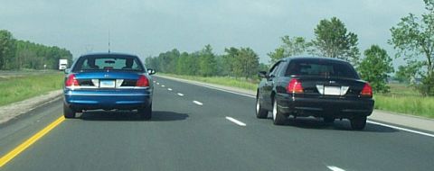 2001 Ford Crown Victoria - Two Ford Crown Victorias, one blue and one black, travel down a highway.