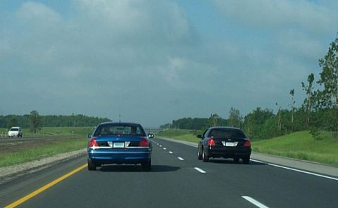 2001 Ford Crown Victoria - Two sedans, one blue and one black, travel down a highway under a cloudy sky.