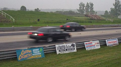 2001 Ford Crown Victoria - Two black sedans race down a track, with various sponsor signs visible in the foreground.