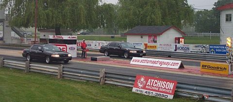 2001 Ford Crown Victoria - Two black Ford Crown Victorias are lined up at the start of a drag race, with a person standing between them.