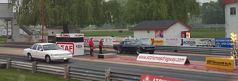 Drag strip with a white sedan and a black coupe at the starting line, with spectators and buildings in the background
