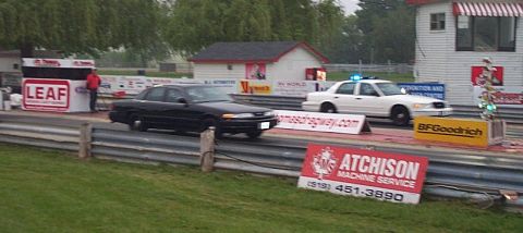 2001 Ford Crown Victoria - Two sedans, one black and one white, are on a drag strip with a building and trees in the background.