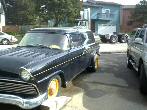 1972 Ford Ranchero - A dark blue classic sedan with yellow wheels is parked on asphalt. A silver SUV is parked to its right, and a trailer is visible in the bac…