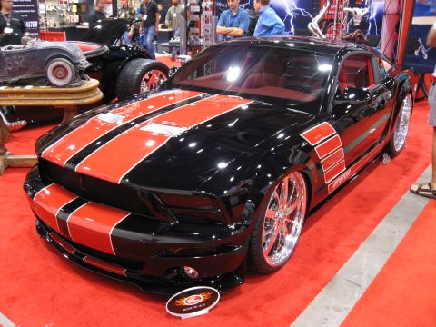 1998 Ford Crown Victoria - A modified black Ford Mustang with red racing stripes is displayed on a red carpet at a car show.