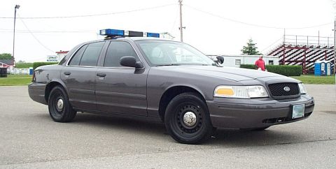 2001 Ford Crown Victoria - A gray police car is parked on pavement with bleachers and portable toilets in the background.