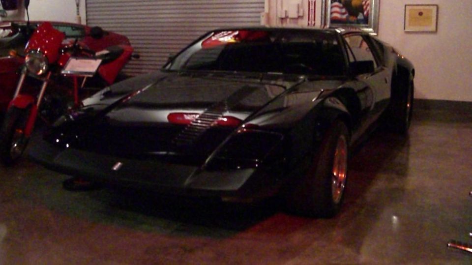 A black wedge-shaped sports car parked indoors next to a red motorcycle
