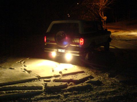 1989 Ford Bronco - The rear of a Ford Bronco is visible at night, with its backup lights on and a spare tire mounted on the tailgate.