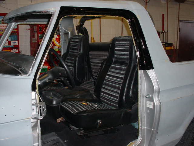Gray 1978 Ford Bronco interior with striped seats and black dashboard, viewed from the driver's side door opening