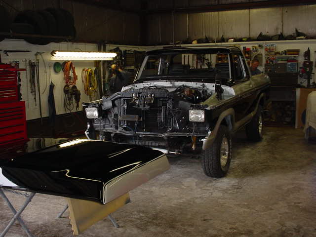 1978 Ford Bronco in garage, hood off, engine exposed. Black hood on stand in foreground