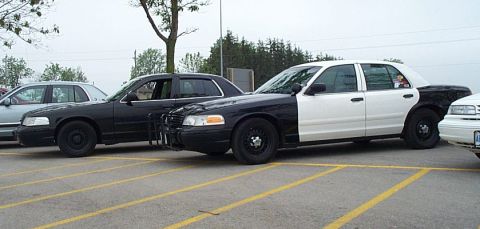2001 Ford Crown Victoria - Two police cruisers are parked in a lot, one with a two-tone paint scheme and the other all black.