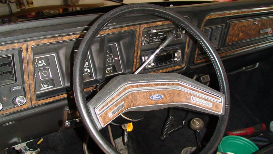 Interior of a vintage Ford vehicle, showing dashboard with wood grain trim, steering wheel with Ford logo, and gauges.