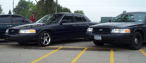2001 Ford Crown Victoria - Three dark Ford Crown Victoria sedans parked side by side in a striped lot on a grey day; the center car is lowered on polished multi-spoke…