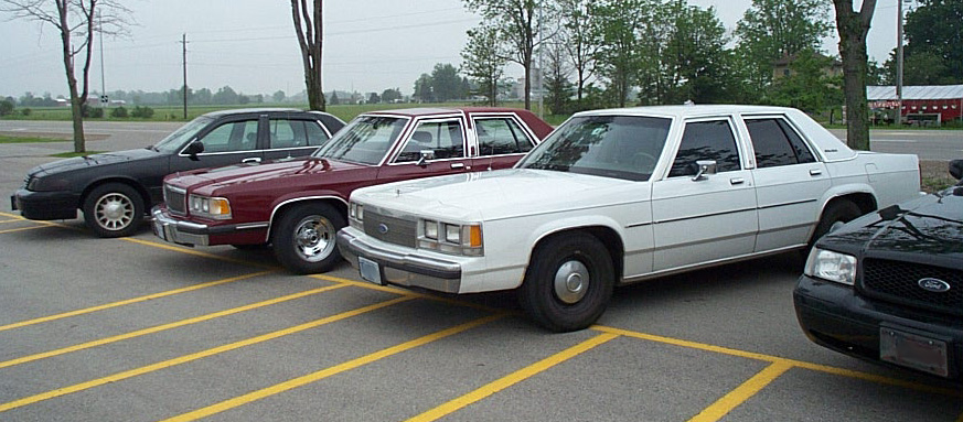 Three sedans parked in a lot, one white, one maroon, and one black. The white car is a Ford Crown Victoria from the 1980s