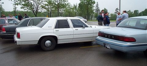 2001 Ford Crown Victoria - A white sedan is parked in a lot with other vehicles and people nearby.