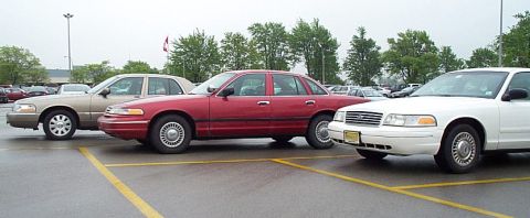 2001 Ford Crown Victoria - Three sedans, including a white and red 2001 Ford Crown Victoria, are parked in a lot on a cloudy day.