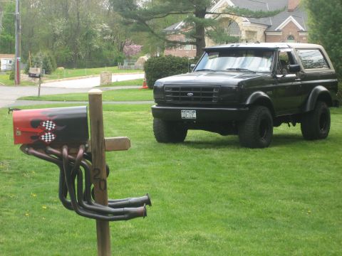 1995 Ford Bronco - A black Ford Bronco is parked on a lawn next to a custom mailbox racing flags and exhaust pipes.