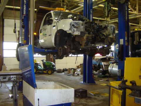 1992 Ford Bronco - A white truck is suspended on a lift with its front end disassembled, undergoing repairs in a workshop. A green lawn mower is parked nearby.