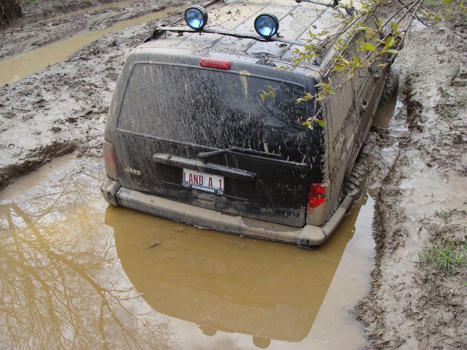 Black SUV stuck in mud, rear view, with 'LAND A 1' license plate and two blue lights on the roof