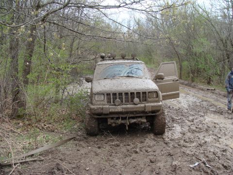 1992 Ford Bronco - Jeep Cherokee stuck deep in mud on a wooded trail, with the driver's door open.