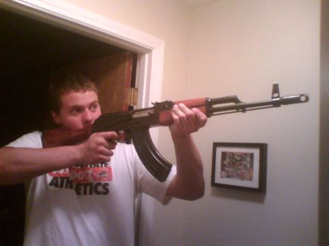 1993 Ford Bronco - A man aims an AK-47 rifle indoors, with a framed picture on the wall.