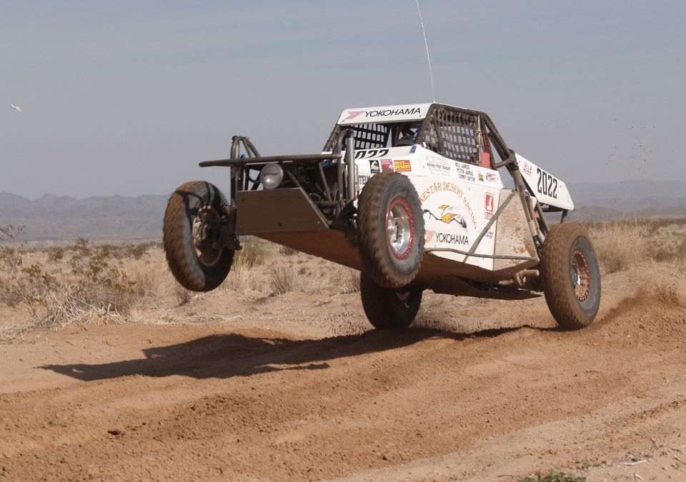 White off-road buggy with number 2022 airborne over a dirt track, desert landscape
