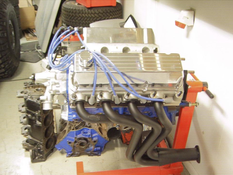 V8 engine on orange stand with aluminum valve covers, black headers, and blue plug wires