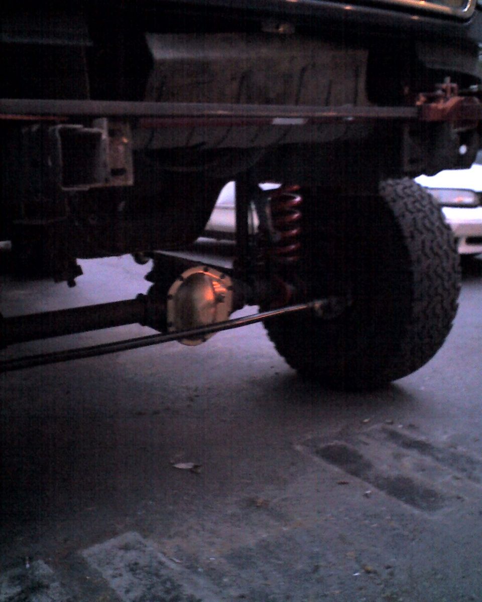 1996 Ford F-150 rear axle, suspension, and tire. Red coil spring visible. Another vehicle is partially seen in the background